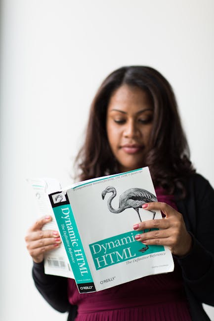 A woman with dark hair, dressed in a black blazer and maroon top, is holding a book titled 'Dynamic HTML' featuring a flamingo illustration on the cover. She is standing indoors against a plain white 